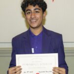 A teenage boy, with dark curly hair and purple blazer smiles at the camera as he holds up his certificate for presenting his speech