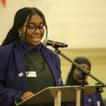 A teen girl with braids, dark glasses and a purple blazer smiles as she stands at the lectern and delivers her speech to the crowd