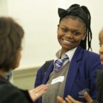 A teenage girls, in purple blazer with braids and a large dark bow in her hair smiles broadly at the camera as a member of the audience talks to her about her speech.