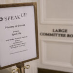 A brown, wooden lectern - featuring a white poster reading 'Speak Up' Ministry of Stories - stands outside a grand, white door with the words 'Large Committee Room' embossed on it