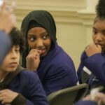 A young pupil with black headscarf and bright purple blazer looks over her shoulder, chewing her nails, looking nervously at a friend.