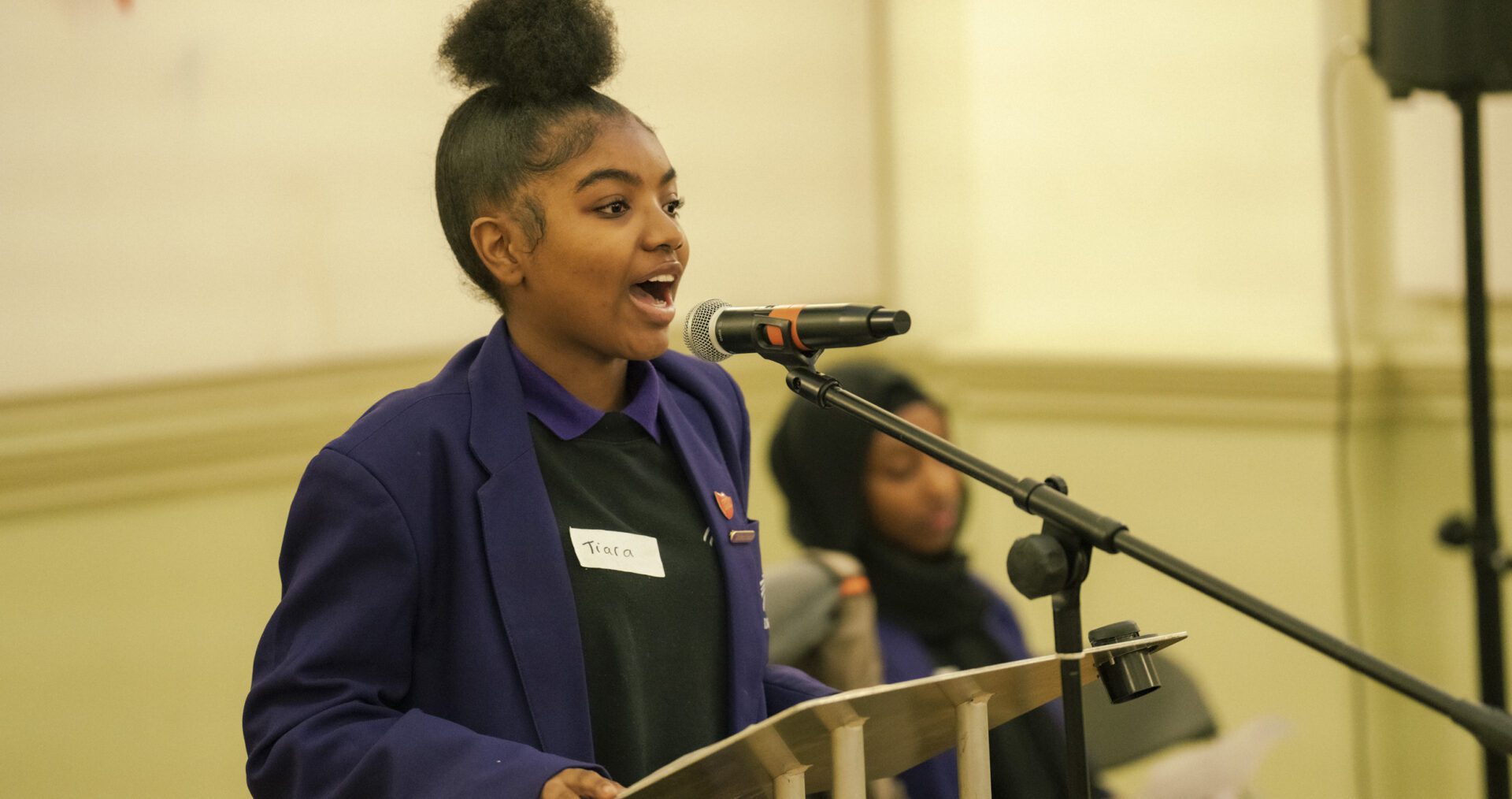 A teen girl with dark hair in a large afro bun on top of her head, wearing a purple blazer, speaks animatedly to the crowd ahead of the lectern she stands in front of