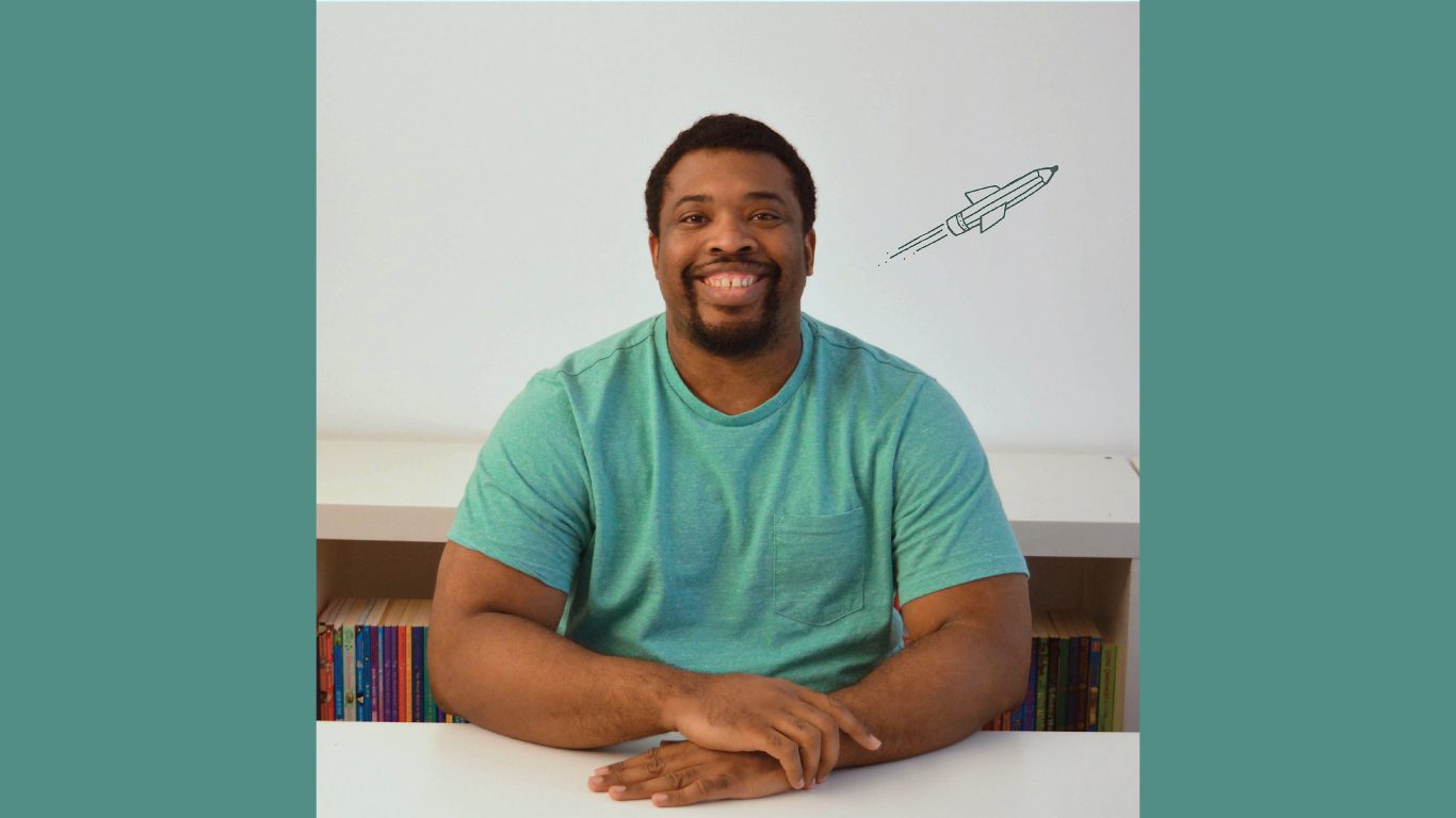 Mayo, a black man with dark hair and a trimmed beard, sits smiling at the camera his hand clasped in front of him. He wears a bright, teal t-shirt and has a small illustrated rocket blasting off on the wall behind him.