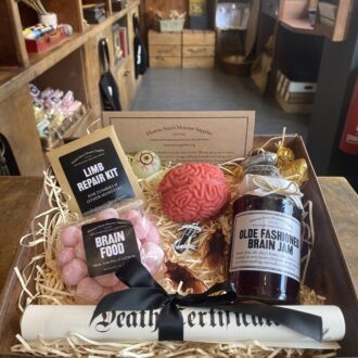 A square hamper packed with straw sits on a wooden counter top. It is full of Monster Supplies goodies including a Death Certificate, Brain Food and Brain Jam. 
