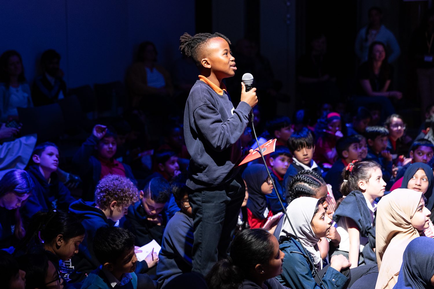 A young black boy stands proudly holding a microphone, addressing someone off camera. He is surrounded by other young children, sitting on the floor of a hall, who are looking up at him.