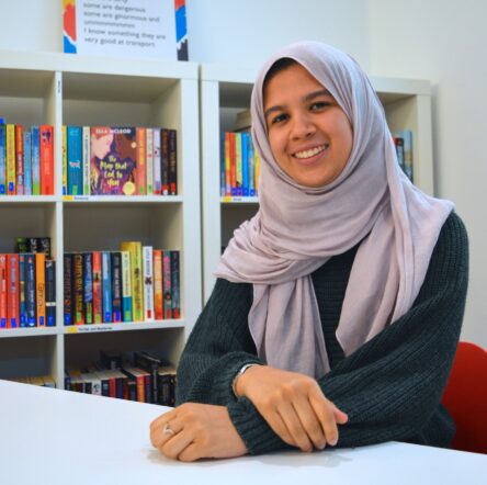 Fathima, a young woman, sits smiling at the camera. She wears a dark, long sleeve top and a pale lilac hijab.