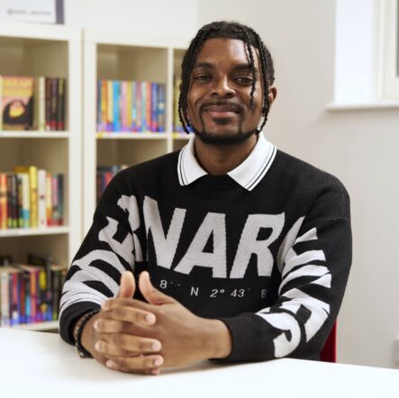 Regan - a black man with dark twists hair - sits with his hands folded in front of him. He wears a dark jumper with white writing on it and a white collar. He smiles happily at the camera.