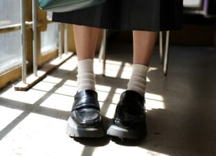 A close up image of a young person wearing short white socks and black school shoes, standing in a classroom with a desk behind them.