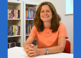 Laura - a white woman with long brown hair - sits smiling in front of a colourful bookshelf. Laura is wearing a peach t-shirt, a short silver necklace and a silver watch.